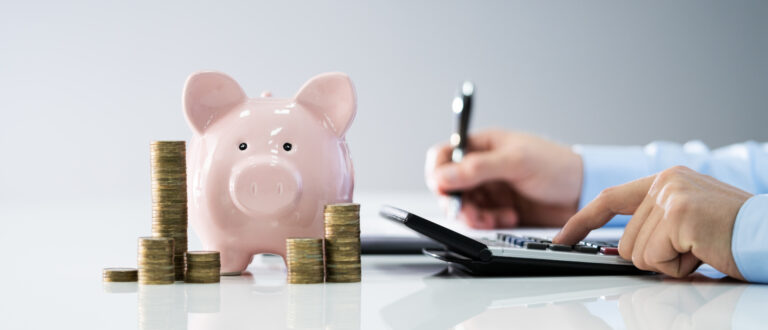 En person räknar på en miniräknare. På bordet står en spargris och en hög med mynt.