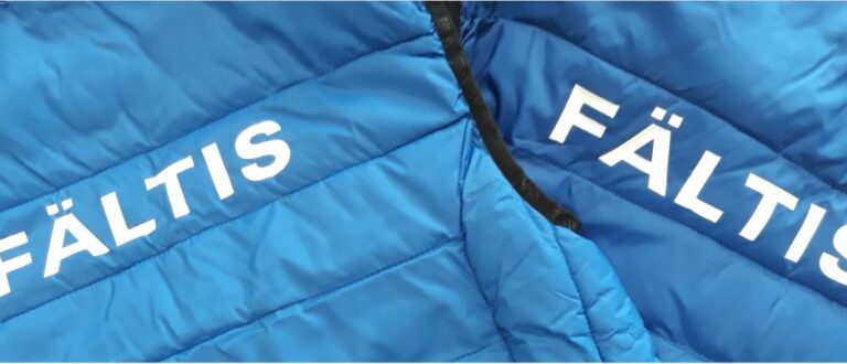 Närbild på två blå vadderade västar där det står Fältis på ryggen i vitt