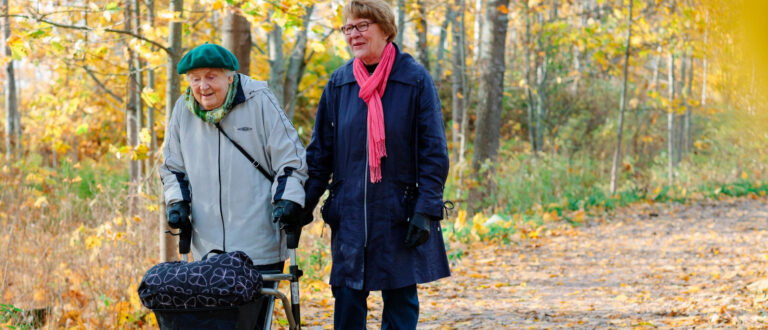 Det är höst. En kvinna leder en äldre kvinna med rullator.