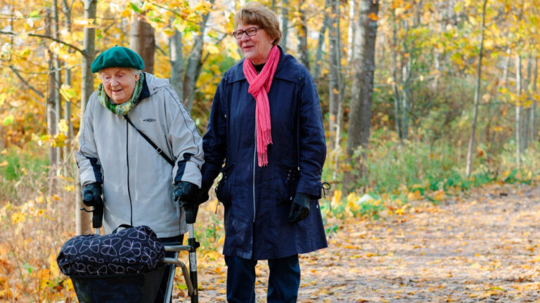 On syksy. Nainen saattaa iäkkäämpää rollaattorin kanssa kulkevaa naista.