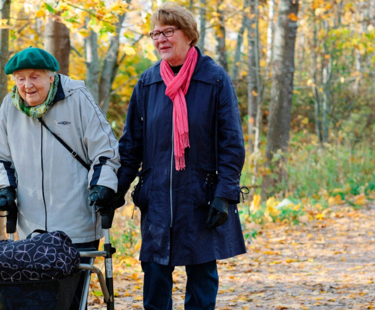 On syksy. Nainen saattaa iäkkäämpää rollaattorin kanssa kulkevaa naista.